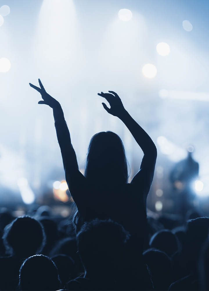 Persona entre el público de un concierto levanta ambos brazos hacia el escenario, apareciendo en silueta por el fuerte contraluz. Al fondo se distinguen focos y luces en tonos azulados, con figuras desenfocadas sobre el escenario y una multitud ocupando la parte inferior de la imagen. La iluminación crea una atmósfera de espectáculo en directo y resalta el contraste entre la figura central y el fondo luminoso.