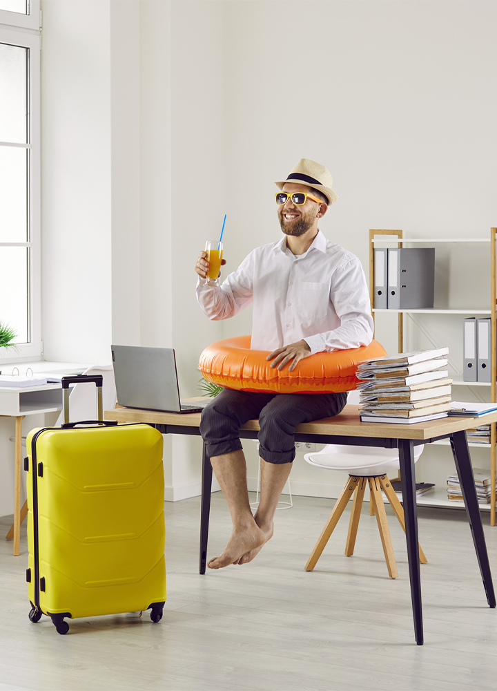 Persona sentada sobre una mesa de oficina con un flotador naranja alrededor de la cintura, sosteniendo una bebida con pajita. Lleva sombrero, gafas de sol y ropa informal, con los pies descalzos. A su lado hay una maleta amarilla, un ordenador portátil abierto y una pila de libros. El espacio es una oficina luminosa con estanterías, archivadores y grandes ventanas, creando una escena que combina trabajo y ambiente vacacional.