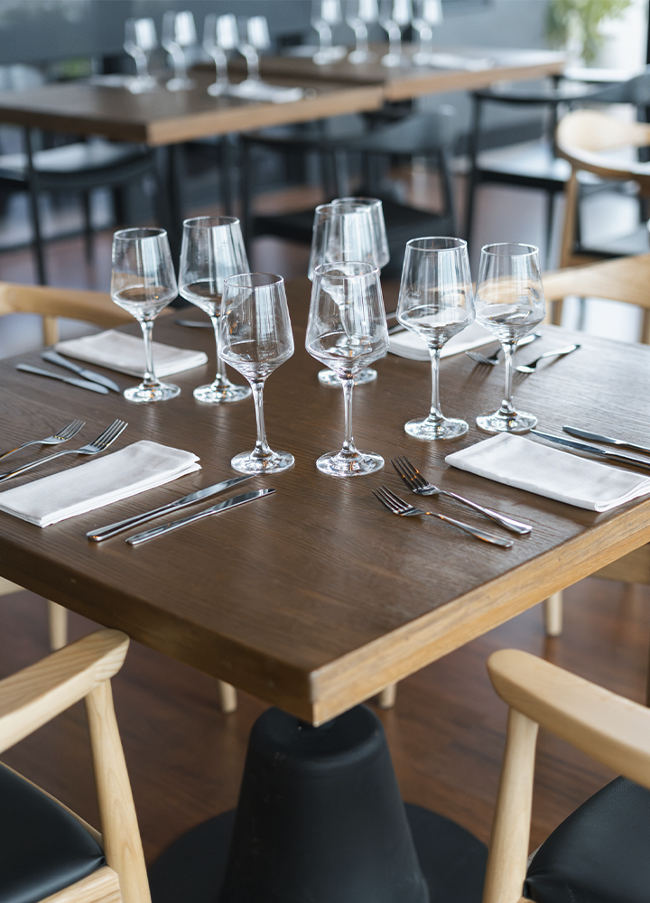 Mesa de restaurante preparada para varias personas, con copas de vino vacías distribuidas en el centro, cubiertos alineados y servilletas blancas dobladas junto a cada cubierto. La mesa de madera oscura está rodeada de sillas de diseño claro, mientras otras mesas preparadas aparecen desenfocadas al fondo. La iluminación suave y el ambiente ordenado transmiten una atmósfera elegante y acogedora.