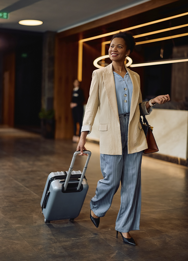 Persona caminando por el vestíbulo de un hotel mientras arrastra una maleta rígida de color gris con ruedas. Lleva una chaqueta clara, pantalones de rayas finas y un bolso colgado del hombro. El espacio presenta una decoración elegante con revestimientos de madera, mostrador de recepción y líneas de iluminación cálida en el fondo. La maleta ocupa el primer plano y la escena transmite una situación de llegada, salida o desplazamiento dentro de un alojamiento.
