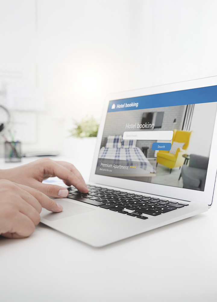 Primer plano de una persona utilizando un ordenador portátil sobre una superficie blanca mientras accede a una página web de reservas de alojamiento. En la pantalla aparece el texto “Hotel booking”, una barra de búsqueda y una fotografía de una habitación. Las manos se apoyan sobre el teclado y el panel táctil del ordenador. El entorno es luminoso, con fondo desenfocado en tonos claros que dirige la atención hacia el proceso de búsqueda y reserva de alojamiento en línea.