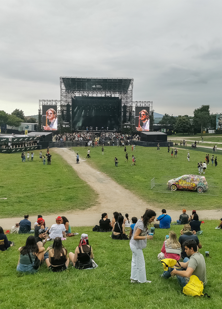 Festival o concierto al aire libre en una amplia explanada de césped. En el centro se alza un gran escenario con dos pantallas gigantes a ambos lados que muestran a la persona que actúa. Decenas de asistentes se distribuyen por el recinto, algunos sentados en la hierba y otros caminando por los senderos que conducen hacia el escenario. En primer plano, varios grupos descansan sobre el césped, mientras a la derecha destaca un coche cubierto de grafitis y colores llamativos. El cielo está nublado y aporta una atmósfera abierta y tranquila a la escena.