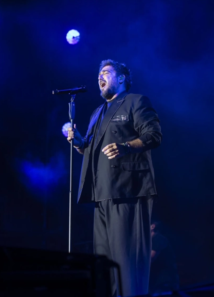 Persona sobre un escenario cantando frente a un micrófono con pie. Viste un conjunto oscuro y aparece iluminada por focos en tonos azules que dominan toda la escena. El fondo permanece oscuro y difuminado, con algunas luces circulares suspendidas que refuerzan el ambiente de concierto en directo.