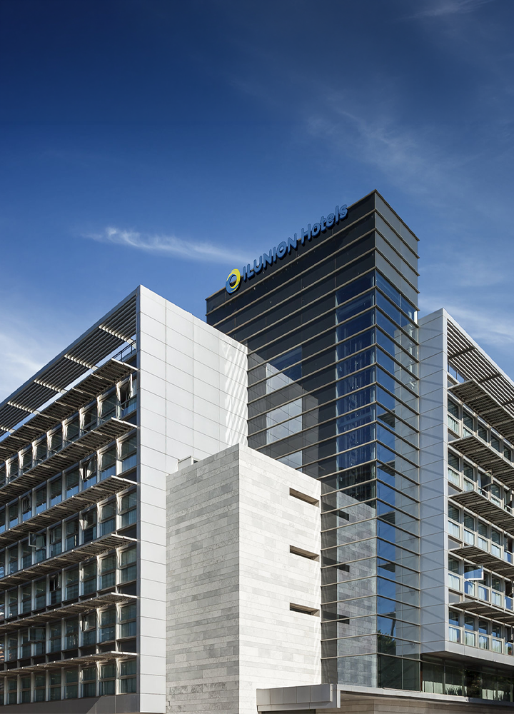 Fachada de un edificio moderno de ILUNION Hotels, con una torre central acristalada y el logotipo de la marca en la parte superior. A ambos lados se extienden alas simétricas con filas de habitaciones y balcones protegidos por lamas metálicas. La arquitectura combina cristal, metal y piedra clara bajo un cielo azul despejado.