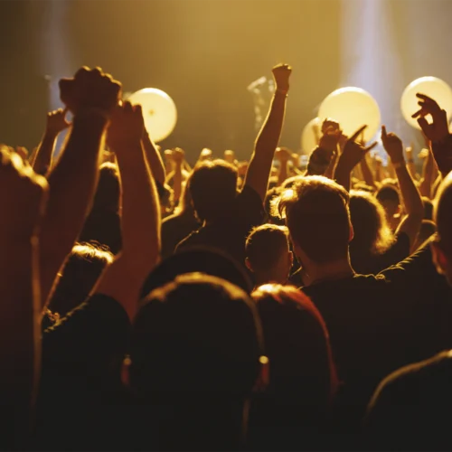Público asistiendo a un concierto o espectáculo en directo, con numerosos brazos levantados hacia el escenario. La escena está iluminada por una intensa luz dorada que crea siluetas y contraluces entre la multitud. Al fondo se distinguen grandes globos claros y focos de escenario difuminados, aportando una atmósfera festiva y multitudinaria.