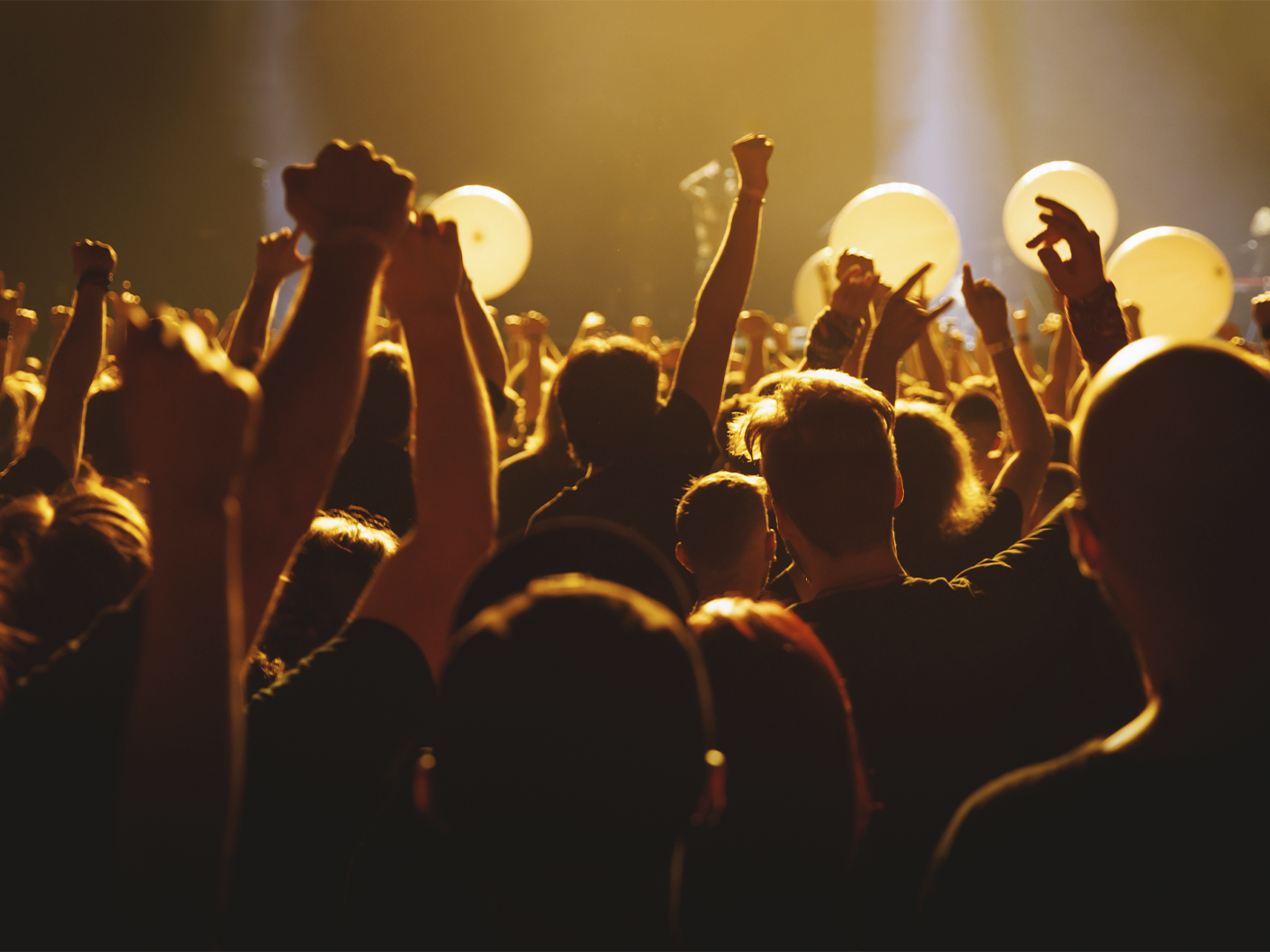Público asistiendo a un concierto o espectáculo en directo, con numerosos brazos levantados hacia el escenario. La escena está iluminada por una intensa luz dorada que crea siluetas y contraluces entre la multitud. Al fondo se distinguen grandes globos claros y focos de escenario difuminados, aportando una atmósfera festiva y multitudinaria.