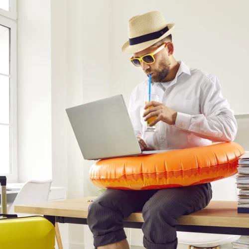 Persona sentada sobre una mesa de oficina utilizando un ordenador portátil mientras lleva un flotador naranja alrededor de la cintura. Sostiene un vaso con bebida y pajita, y viste sombrero y gafas de sol. A la izquierda hay una maleta amarilla de viaje y, a la derecha, una pila de documentos. El entorno es una oficina luminosa con grandes ventanales, estanterías y mobiliario de trabajo, creando un contraste entre el ambiente laboral y elementos asociados a las vacaciones.