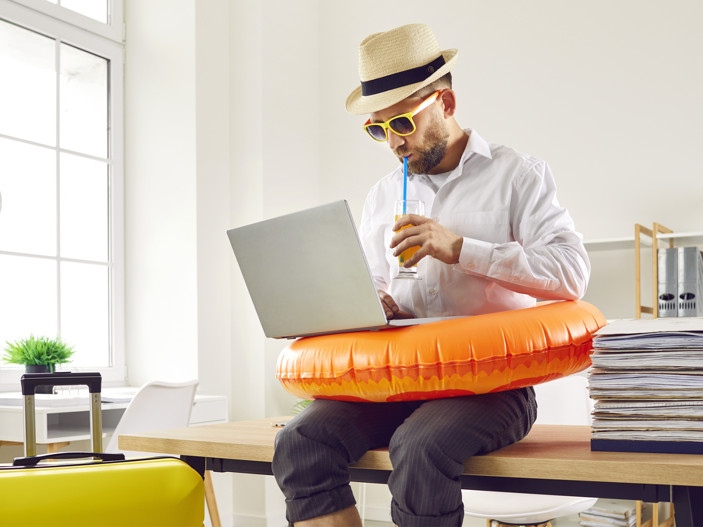 Persona sentada sobre una mesa de oficina utilizando un ordenador portátil mientras lleva un flotador naranja alrededor de la cintura. Sostiene un vaso con bebida y pajita, y viste sombrero y gafas de sol. A la izquierda hay una maleta amarilla de viaje y, a la derecha, una pila de documentos. El entorno es una oficina luminosa con grandes ventanales, estanterías y mobiliario de trabajo, creando un contraste entre el ambiente laboral y elementos asociados a las vacaciones.
