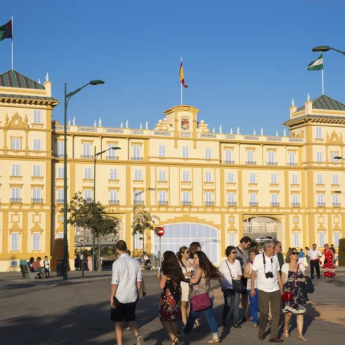 Edificio histórico de gran tamaño con fachada en tonos crema y amarillo, decorada con molduras y torres en ambos extremos. En la parte central ondea una bandera de España y, sobre las torres laterales, otras banderas. Frente al edificio, varias personas cruzan una amplia plaza o avenida peatonal bajo la luz cálida del sol. El cielo aparece completamente despejado y el entorno incluye farolas, árboles y zonas de paso alrededor del inmueble.