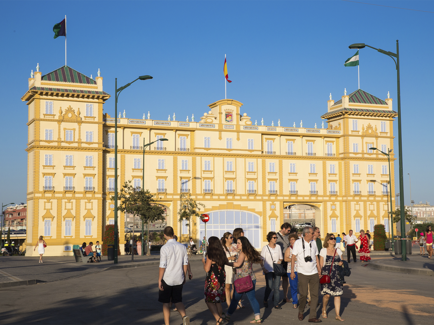 Edificio histórico de gran tamaño con fachada en tonos crema y amarillo, decorada con molduras y torres en ambos extremos. En la parte central ondea una bandera de España y, sobre las torres laterales, otras banderas. Frente al edificio, varias personas cruzan una amplia plaza o avenida peatonal bajo la luz cálida del sol. El cielo aparece completamente despejado y el entorno incluye farolas, árboles y zonas de paso alrededor del inmueble.