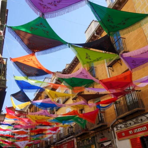 Calle estrecha decorada con coloridos mantones bordados suspendidos entre los edificios, creando un techo multicolor sobre la vía. Los tejidos en tonos rosa, verde, azul, amarillo, naranja, rojo y negro cuelgan formando franjas paralelas bajo un cielo azul despejado. A ambos lados se alinean fachadas tradicionales con balcones de hierro forjado, comercios y faroles, mientras al fondo la calle continúa bajo la sucesión de adornos festivos.