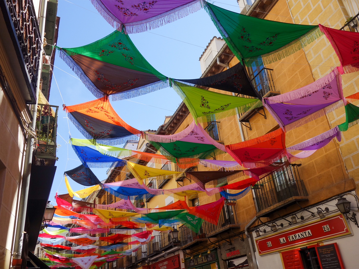 Calle estrecha decorada con coloridos mantones bordados suspendidos entre los edificios, creando un techo multicolor sobre la vía. Los tejidos en tonos rosa, verde, azul, amarillo, naranja, rojo y negro cuelgan formando franjas paralelas bajo un cielo azul despejado. A ambos lados se alinean fachadas tradicionales con balcones de hierro forjado, comercios y faroles, mientras al fondo la calle continúa bajo la sucesión de adornos festivos.