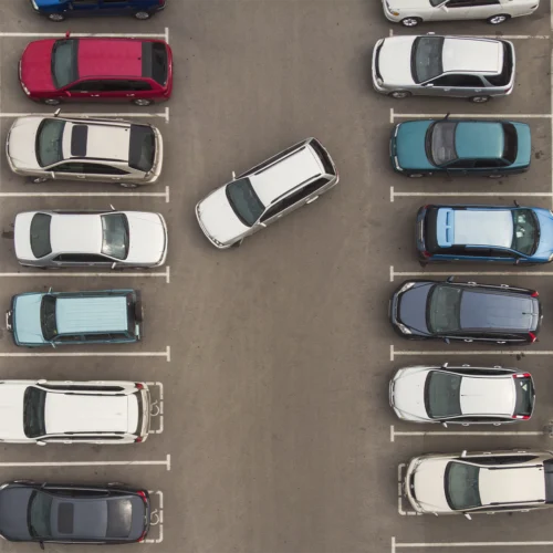 Vista aérea de un aparcamiento con vehículos estacionados en filas ordenadas a ambos lados. En el centro, un coche blanco maniobra en diagonal entre las plazas, contrastando con el resto de vehículos aparcados. Las marcas blancas delimitan claramente cada espacio sobre el asfalto gris, creando una composición simétrica vista desde arriba.