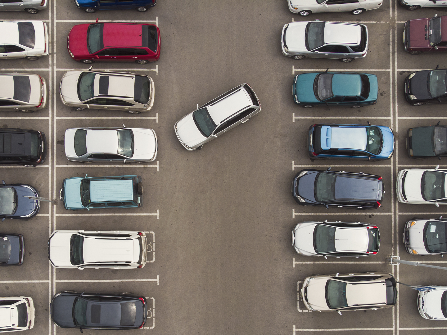 Vista aérea de un aparcamiento con vehículos estacionados en filas ordenadas a ambos lados. En el centro, un coche blanco maniobra en diagonal entre las plazas, contrastando con el resto de vehículos aparcados. Las marcas blancas delimitan claramente cada espacio sobre el asfalto gris, creando una composición simétrica vista desde arriba.