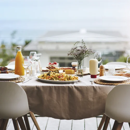 Mesa preparada para una comida en una terraza con vistas al mar. Sobre un mantel beige hay platos, copas, cubiertos, una ensalada de pasta, pan, quesos, salsas, una botella de zumo y un pequeño jarrón con flores. Dos sillas blancas ocupan el primer plano, mientras el fondo aparece suavemente desenfocado con el horizonte marítimo visible tras una barandilla de cristal.