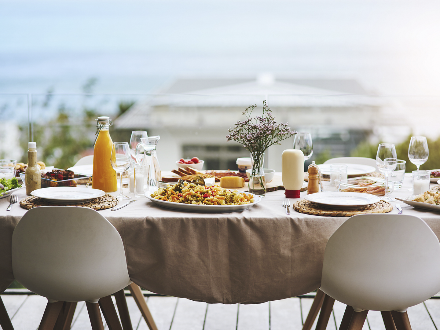 Mesa preparada para una comida en una terraza con vistas al mar. Sobre un mantel beige hay platos, copas, cubiertos, una ensalada de pasta, pan, quesos, salsas, una botella de zumo y un pequeño jarrón con flores. Dos sillas blancas ocupan el primer plano, mientras el fondo aparece suavemente desenfocado con el horizonte marítimo visible tras una barandilla de cristal.