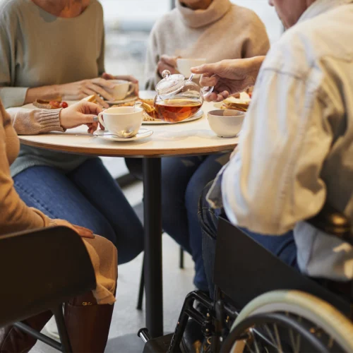 Personas reunidas alrededor de una mesa compartiendo una merienda o pausa para el té. En el centro, una mano sirve té desde una tetera de cristal transparente hacia una taza blanca. Sobre la mesa hay varias tazas, platos con comida y pequeños aperitivos. Una de las personas utiliza una silla de ruedas, visible en el lateral derecho de la imagen. La escena transcurre en un espacio interior con iluminación cálida y un ambiente de encuentro y convivencia.