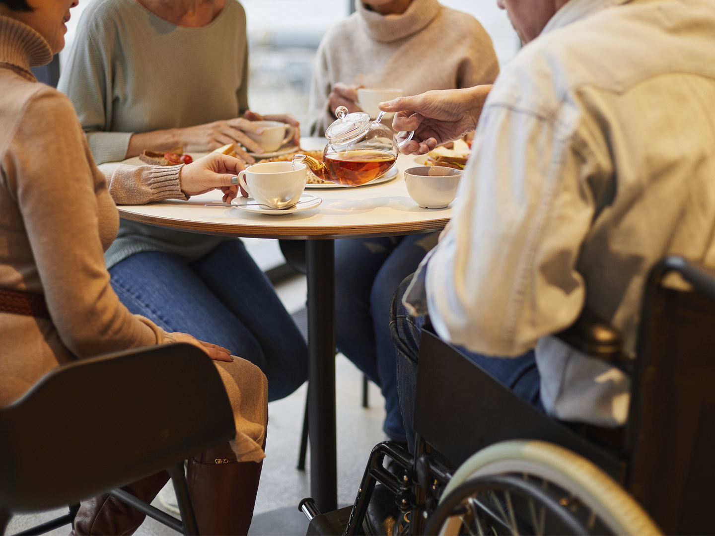 Personas reunidas alrededor de una mesa compartiendo una merienda o pausa para el té. En el centro, una mano sirve té desde una tetera de cristal transparente hacia una taza blanca. Sobre la mesa hay varias tazas, platos con comida y pequeños aperitivos. Una de las personas utiliza una silla de ruedas, visible en el lateral derecho de la imagen. La escena transcurre en un espacio interior con iluminación cálida y un ambiente de encuentro y convivencia.