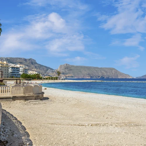 Paseo marítimo junto a una amplia playa de cantos claros y aguas azul turquesa. A la izquierda se observa una barandilla curva y varias palmeras alineadas frente a edificios residenciales de baja altura. Al fondo destacan montañas y promontorios rocosos que se adentran en el mar, bajo un cielo azul con nubes dispersas. La escena combina elementos urbanos y naturales en un entorno costero luminoso y despejado.
