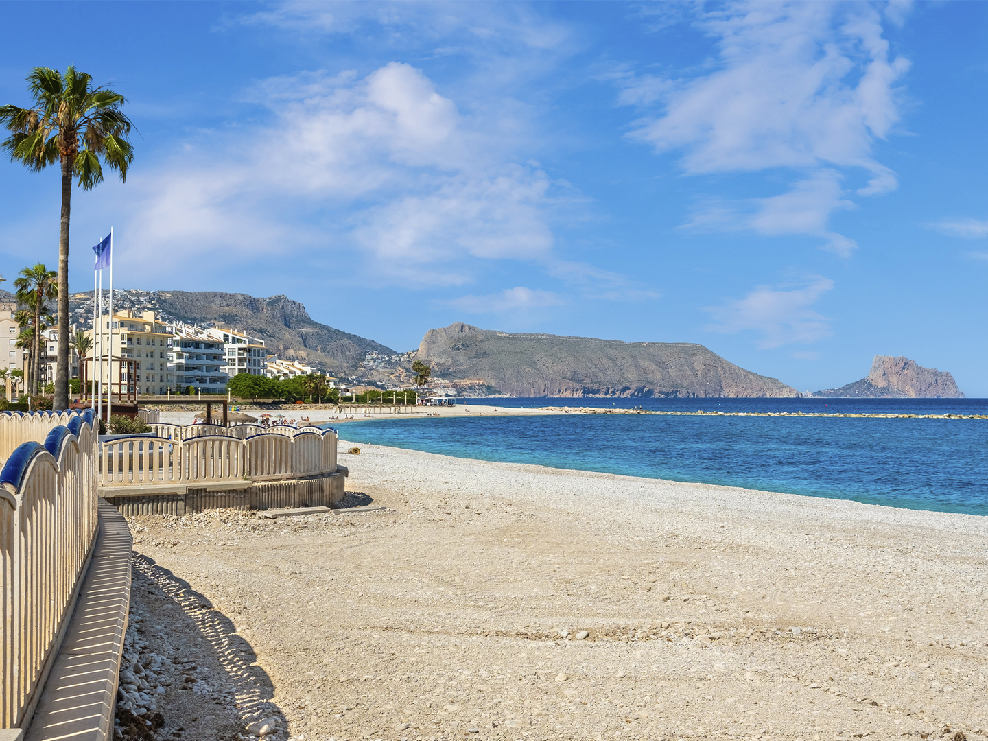 Paseo marítimo junto a una amplia playa de cantos claros y aguas azul turquesa. A la izquierda se observa una barandilla curva y varias palmeras alineadas frente a edificios residenciales de baja altura. Al fondo destacan montañas y promontorios rocosos que se adentran en el mar, bajo un cielo azul con nubes dispersas. La escena combina elementos urbanos y naturales en un entorno costero luminoso y despejado.