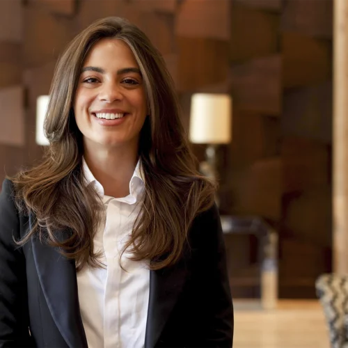 Persona con traje oscuro y camisa blanca posando frente a la cámara en un espacio interior de estilo elegante. Al fondo se observan paneles de madera decorativos, lámparas desenfocadas y mobiliario en tonos neutros. La composición está centrada en la persona, mientras la iluminación cálida y el fondo suavemente desenfocado crean un ambiente propio de la recepción o zona común de un hotel o establecimiento de servicios.