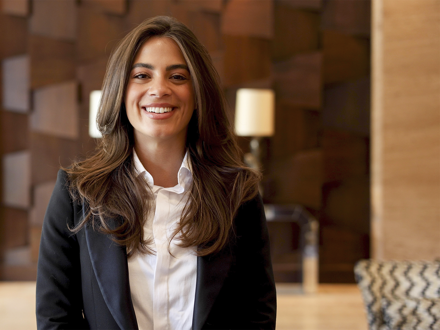 Persona con traje oscuro y camisa blanca posando frente a la cámara en un espacio interior de estilo elegante. Al fondo se observan paneles de madera decorativos, lámparas desenfocadas y mobiliario en tonos neutros. La composición está centrada en la persona, mientras la iluminación cálida y el fondo suavemente desenfocado crean un ambiente propio de la recepción o zona común de un hotel o establecimiento de servicios.