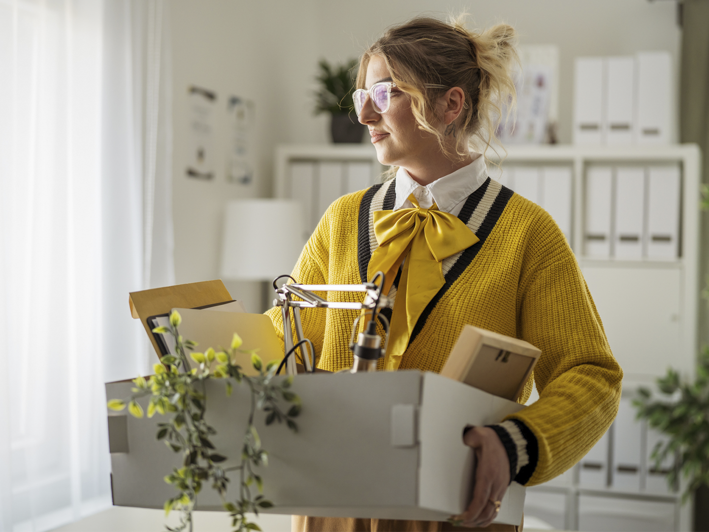 Persona con gafas sostiene una caja con documentos, una lámpara de escritorio y otros objetos de oficina en un espacio de trabajo luminoso. Viste un cárdigan amarillo sobre una camisa blanca con un lazo del mismo color y se encuentra junto a una ventana que deja entrar luz natural. Al fondo se observan estanterías con archivadores, plantas decorativas y mobiliario de oficina en tonos claros.