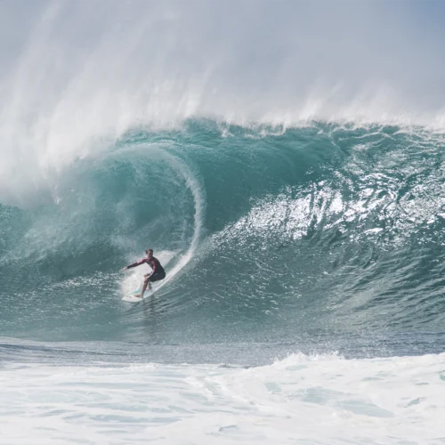 Persona practicando surf sobre una gran ola azul verdosa. La tabla avanza dentro del tubo de la ola mientras el agua se curva formando una cavidad y proyecta una nube de espuma. En primer plano se observa el mar agitado y, al fondo, la cresta de la ola rompe con fuerza bajo una luz brillante.