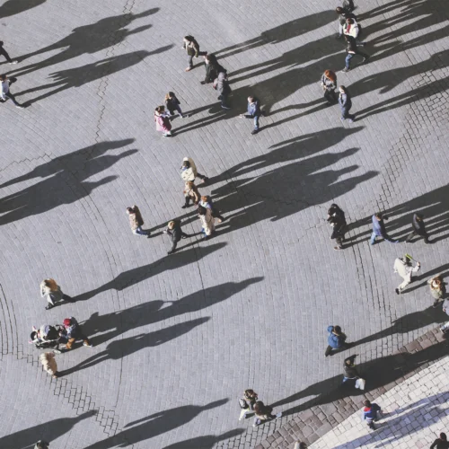 Alt text: Vista aérea de una plaza pavimentada con adoquines claros, recorrida por numerosas personas que caminan en distintas direcciones. La luz baja proyecta sombras muy largas y definidas que se extienden por gran parte del suelo, creando patrones diagonales. Los grupos y transeúntes aparecen dispersos por el espacio abierto, algunos solos y otros acompañados, mientras las sombras ocupan más superficie que las propias figuras. La escena destaca por el contraste entre el pavimento gris claro y las sombras oscuras.