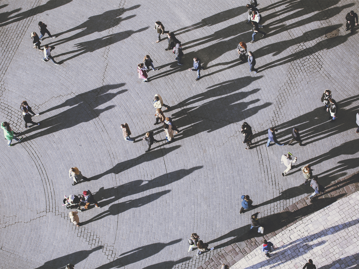 Alt text: Vista aérea de una plaza pavimentada con adoquines claros, recorrida por numerosas personas que caminan en distintas direcciones. La luz baja proyecta sombras muy largas y definidas que se extienden por gran parte del suelo, creando patrones diagonales. Los grupos y transeúntes aparecen dispersos por el espacio abierto, algunos solos y otros acompañados, mientras las sombras ocupan más superficie que las propias figuras. La escena destaca por el contraste entre el pavimento gris claro y las sombras oscuras.