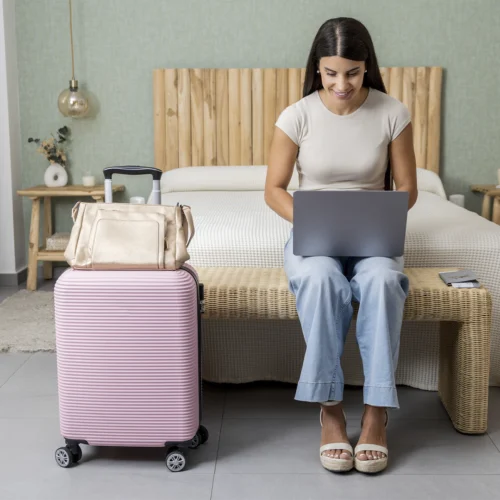Persona sentada en un banco a los pies de una cama, utilizando un ordenador portátil en una habitación de hotel. Frente al banco hay una maleta rígida rosa con ruedas y una bolsa de viaje clara colocada encima. Sobre el asiento, junto a la persona, se observan un pasaporte y tarjetas. La estancia presenta una decoración luminosa con cabecero de madera, mesillas a ambos lados de la cama, lámparas colgantes y ropa de cama en tonos neutros. La escena transmite la planificación o gestión de un viaje desde el alojamiento.