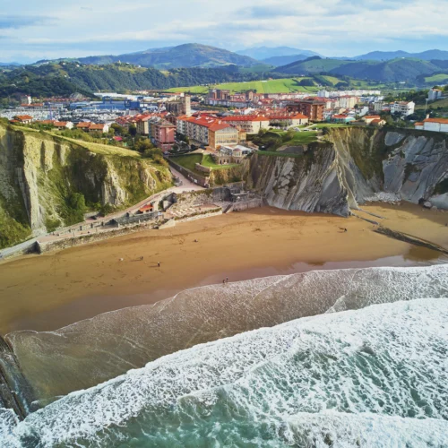Vista aérea de una playa rodeada por espectaculares acantilados de roca estratificada junto a una localidad costera. Las olas rompen sobre la arena mientras un paseo marítimo recorre la base de los acantilados. Al fondo se extiende el núcleo urbano con edificios de tejados rojizos, un puerto y colinas verdes que se elevan hasta las montañas del horizonte bajo un cielo parcialmente nublado.