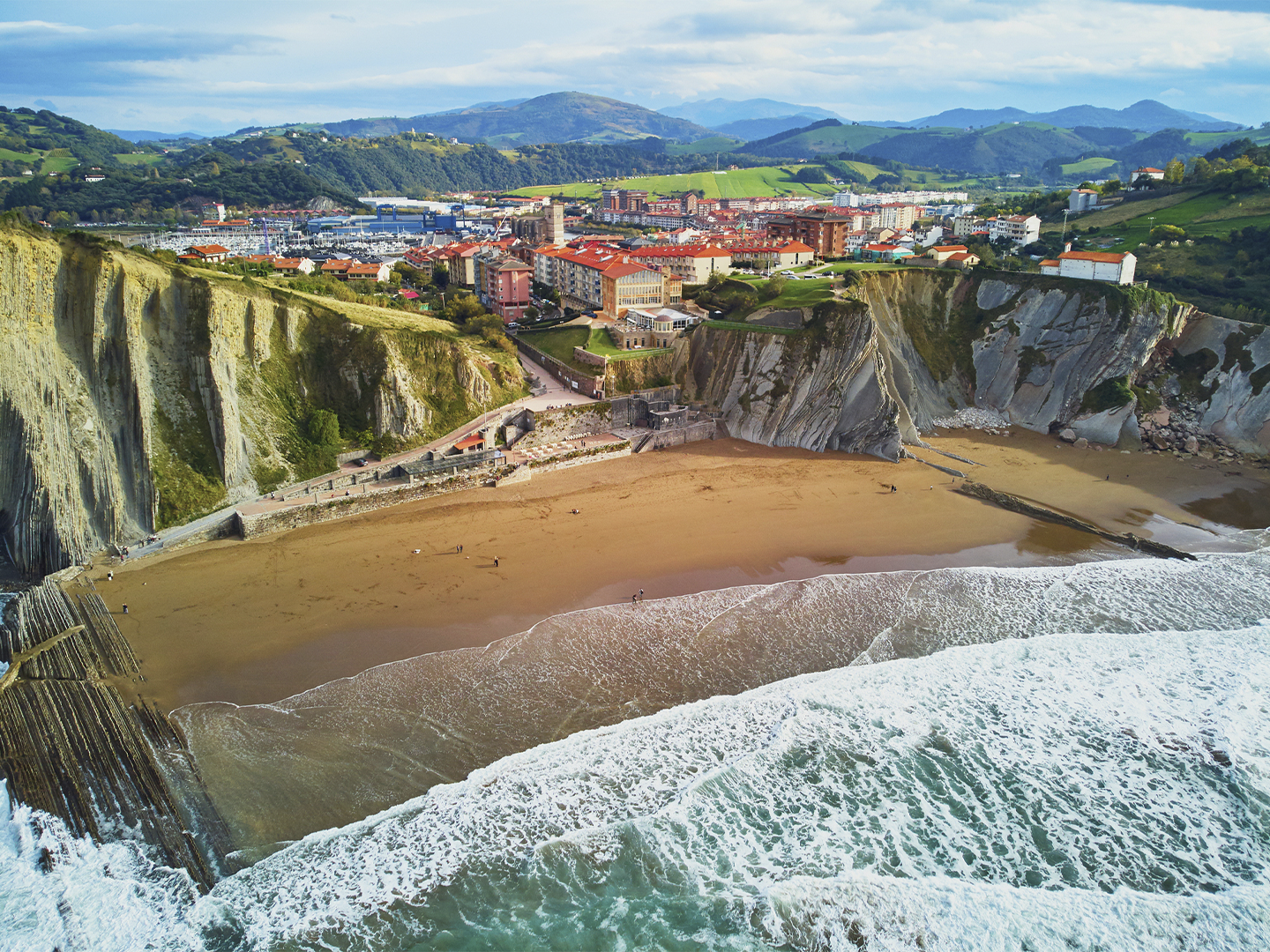 Vista aérea de una playa rodeada por espectaculares acantilados de roca estratificada junto a una localidad costera. Las olas rompen sobre la arena mientras un paseo marítimo recorre la base de los acantilados. Al fondo se extiende el núcleo urbano con edificios de tejados rojizos, un puerto y colinas verdes que se elevan hasta las montañas del horizonte bajo un cielo parcialmente nublado.