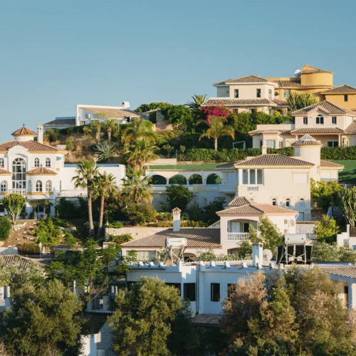 Urbanización residencial en una ladera, con varias viviendas de estilo mediterráneo en tonos claros y tejados de teja. Las casas están rodeadas de palmeras, jardines y abundante vegetación, distribuidas en distintos niveles del terreno. La luz del sol resalta las fachadas y el paisaje bajo un cielo azul despejado.