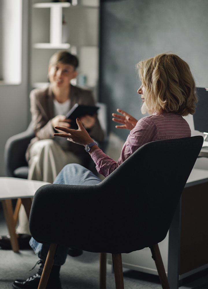 Dos personas mantienen una conversación en un despacho o sala de reunión. En primer plano, una persona sentada en un sillón oscuro gesticula con las manos mientras habla. Frente a ella, otra persona desenfocada sostiene una tableta o libreta para tomar notas. El espacio cuenta con mobiliario moderno, una mesa baja y luz natural que entra desde una ventana lateral, creando un ambiente profesional y tranquilo.