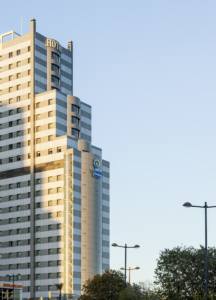 Fachada de un hotel ILUNION de gran altura, con diseño moderno en tonos gris y blanco y numerosas filas de ventanas. En la parte central destaca el logotipo de ILUNION Hotels sobre una estructura curva revestida de metal y cristal. En primer plano aparecen farolas y copas de árboles, mientras la luz cálida del sol ilumina el edificio bajo un cielo completamente despejado.