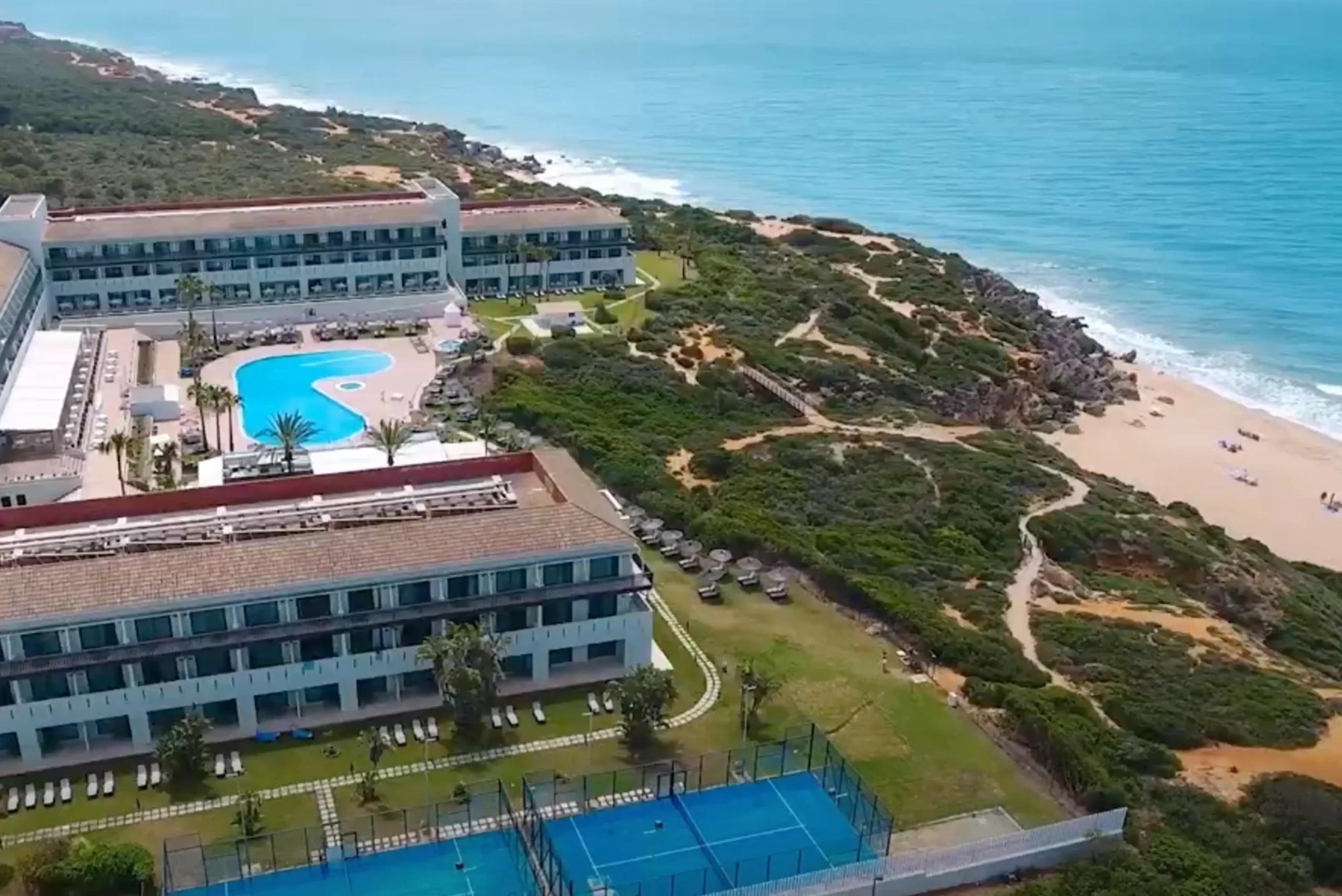 Vista aérea de un complejo hotelero junto a la costa, con varios edificios, una gran piscina de forma orgánica, pistas deportivas y zonas ajardinadas. El establecimiento se encuentra rodeado de vegetación y senderos que descienden hacia una playa de arena bañada por el mar.