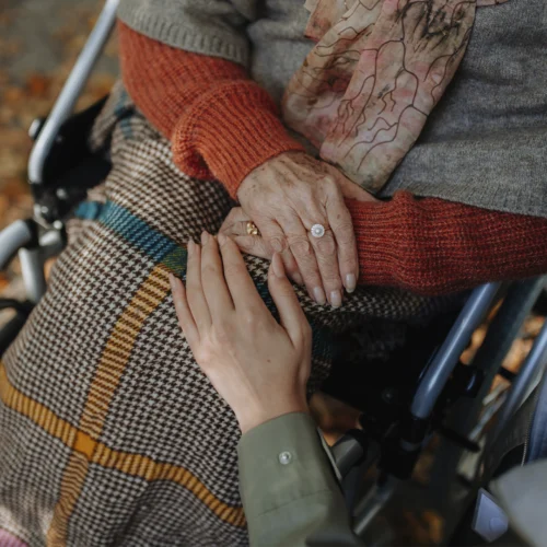 Primer plano de una persona sentada en una silla de ruedas con una manta de cuadros sobre las piernas y las manos apoyadas sobre el regazo. Otra persona acerca una mano en gesto de acompañamiento y apoyo. La escena destaca el contacto entre ambas manos, mientras se aprecian prendas de abrigo en tonos cálidos y un entorno exterior desenfocado al fondo.