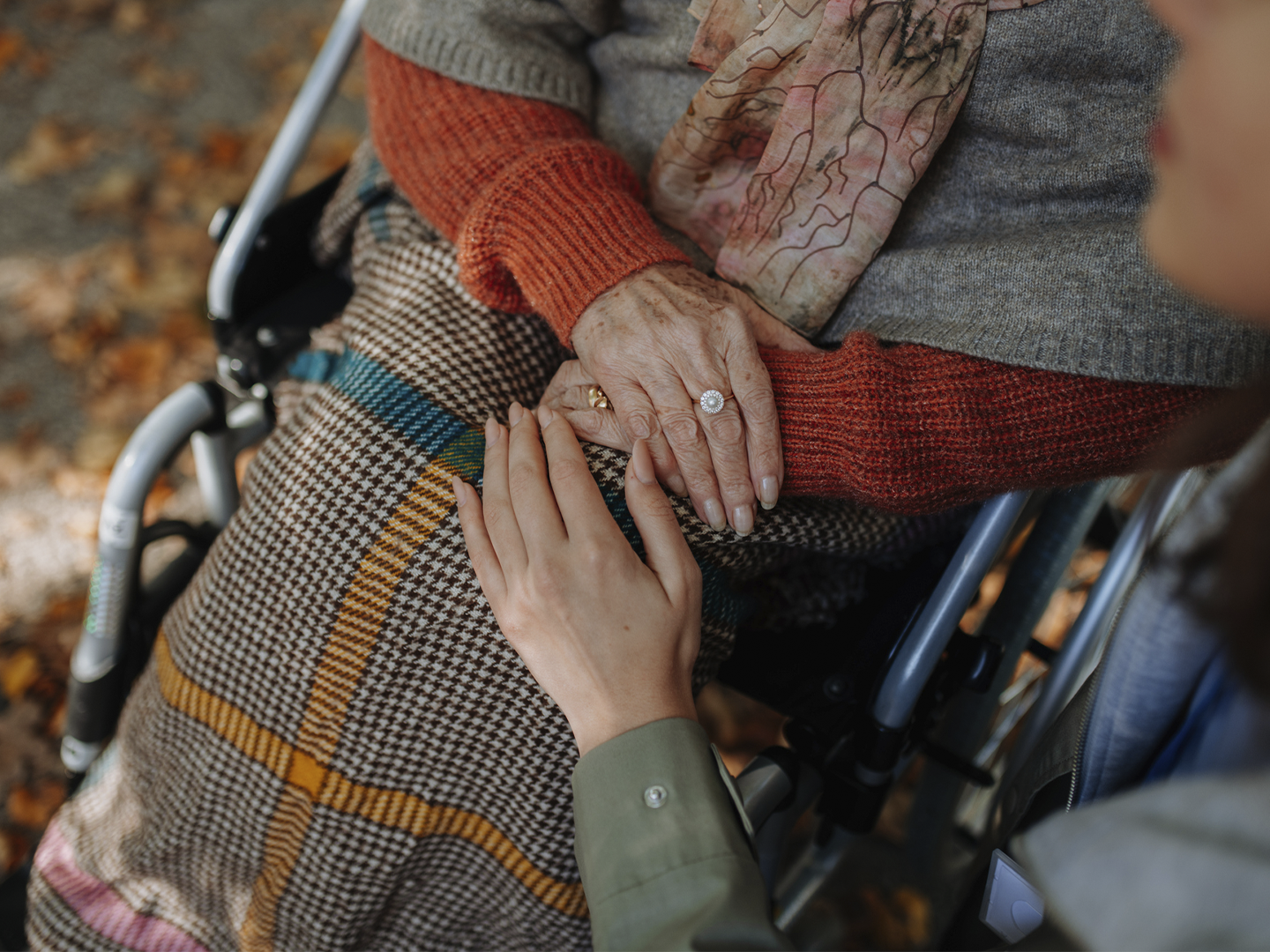 Primer plano de una persona sentada en una silla de ruedas con una manta de cuadros sobre las piernas y las manos apoyadas sobre el regazo. Otra persona acerca una mano en gesto de acompañamiento y apoyo. La escena destaca el contacto entre ambas manos, mientras se aprecian prendas de abrigo en tonos cálidos y un entorno exterior desenfocado al fondo.