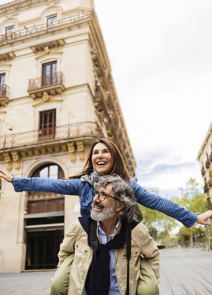 Dos personas en una plaza urbana, una de ellas subida a la espalda de la otra con los brazos extendidos. Al fondo se alza la fachada de un edificio histórico con balcones y detalles ornamentales, mientras la escena transcurre en una calle amplia y despejada con árboles visibles a un lado.