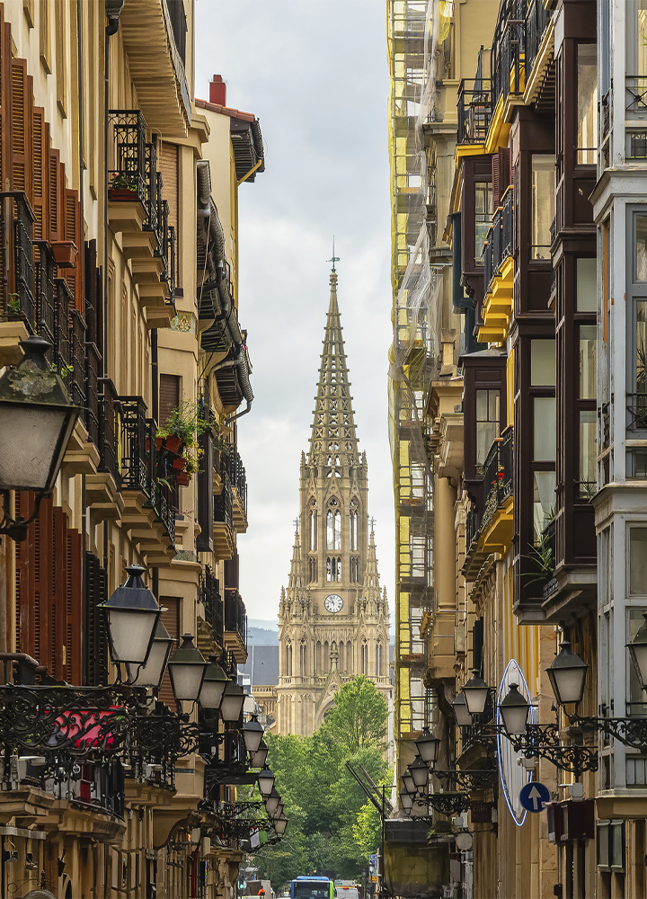 Calle estrecha flanqueada por edificios con balcones y farolas ornamentales, que conduce visualmente hasta la aguja neogótica de una iglesia situada al fondo. La torre, con un reloj visible en su parte central, se eleva sobre una hilera de árboles, mientras las fachadas de ambos lados enmarcan la perspectiva urbana bajo un cielo nublado.