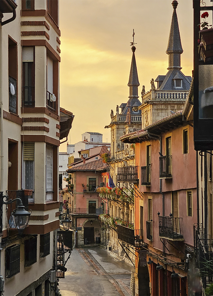 Calle estrecha de un casco histórico con edificios de varios pisos, balcones de hierro forjado y fachadas en tonos claros y ocres. Al fondo sobresalen las torres puntiagudas de un edificio de arquitectura monumental, mientras la calle desciende hacia un arco que conecta varias construcciones. La escena está iluminada por la luz cálida del atardecer, que resalta los detalles arquitectónicos y las macetas con flores en algunos balcones.