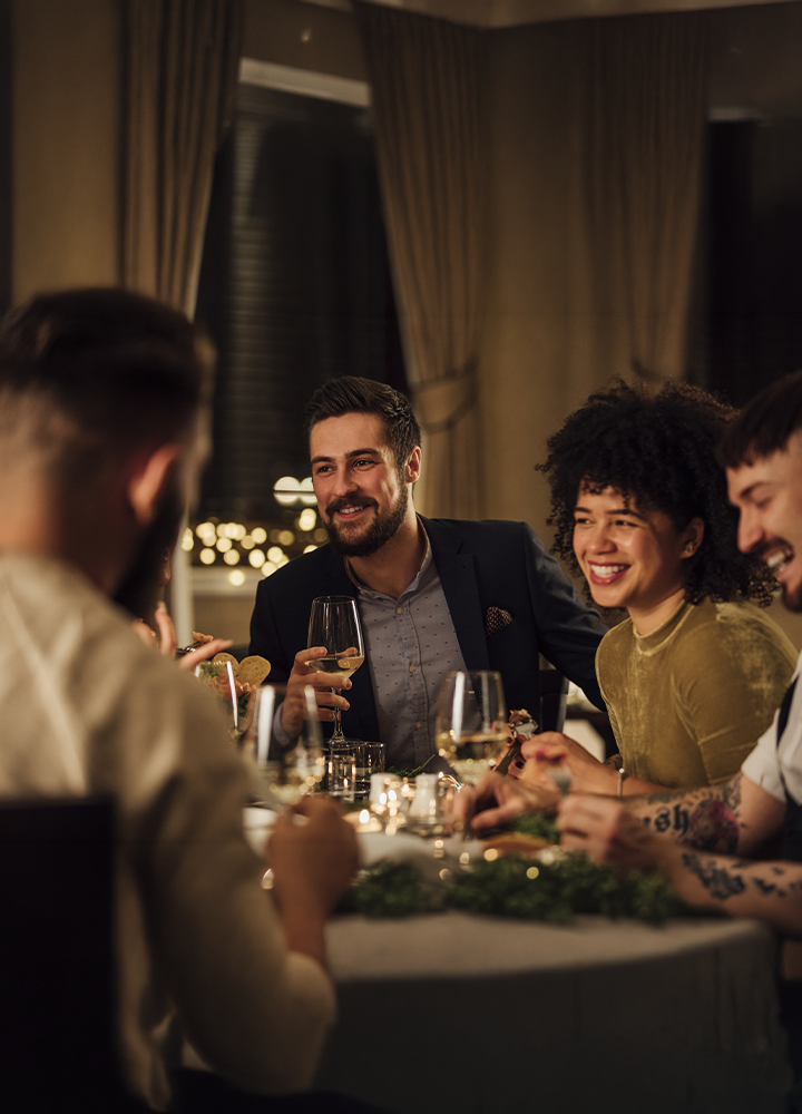 Grupo de personas sentadas alrededor de una mesa en un restaurante o salón con iluminación cálida. Varias copas y platos ocupan el centro de la mesa mientras las personas conversan. Al fondo se ven grandes cortinas y luces desenfocadas que crean un ambiente acogedor e íntimo.