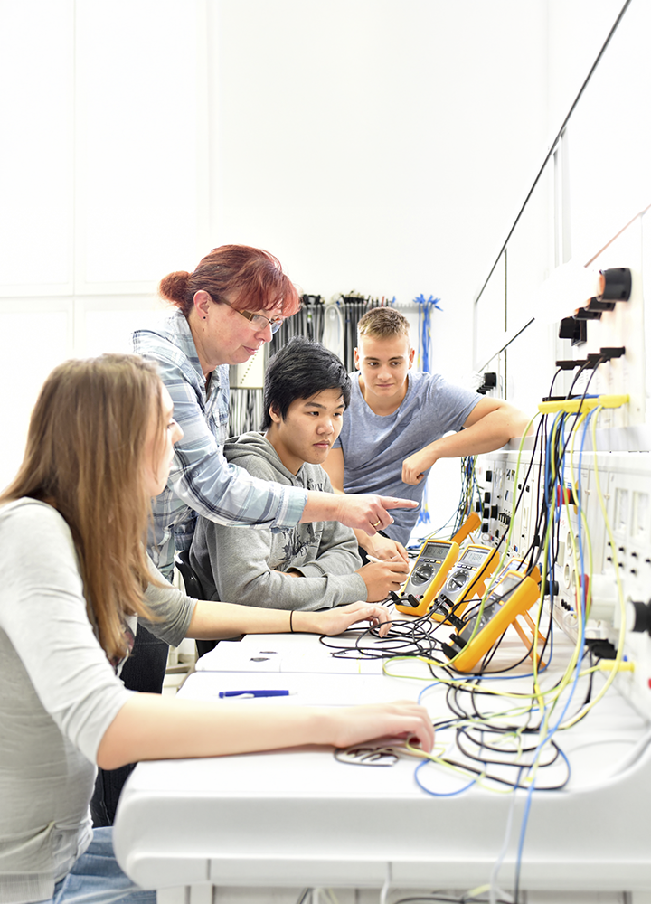 Grupo de personas trabaja en un laboratorio de electrónica equipado con paneles de conexiones, cables y equipos de medición. En primer plano, una persona señala un panel mientras otras observan y manipulan instrumentos de prueba conectados a distintos circuitos. Sobre las mesas se distribuyen multímetros, cables de colores y material técnico utilizado para prácticas de formación en electricidad o electrónica.