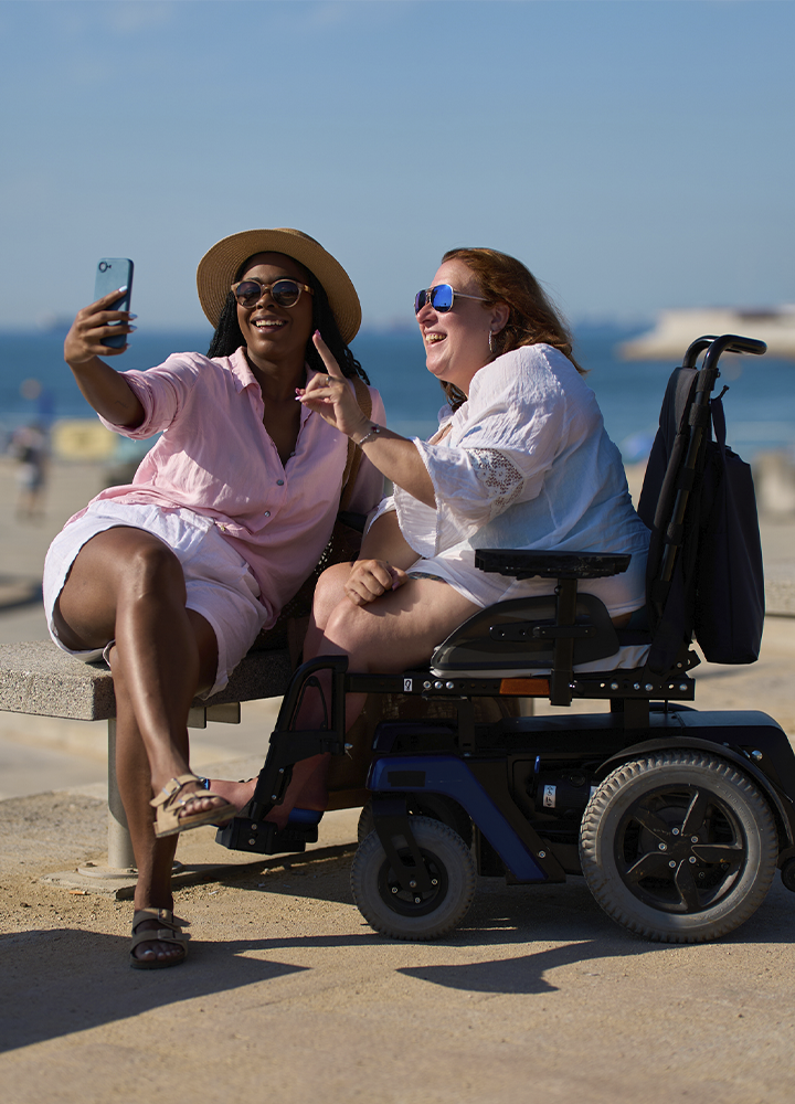 Dos personas posan para hacerse un selfi junto al mar. Una sostiene un teléfono móvil mientras ambas aparecen sentadas en un banco. A la derecha se ve una silla de ruedas eléctrica. El fondo muestra una playa y el agua bajo un cielo despejado.