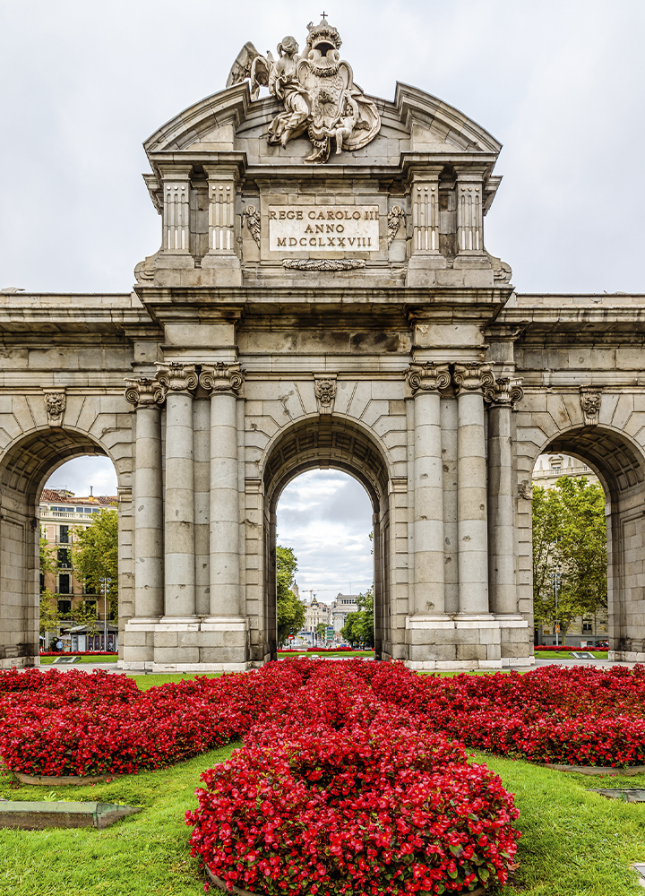 Puerta monumental de piedra con tres arcos, rodeada por amplios parterres de flores rojas y césped verde. La estructura de estilo clásico presenta columnas, esculturas ornamentales y una inscripción en la parte superior. A través del arco central se aprecia una avenida arbolada al fondo.