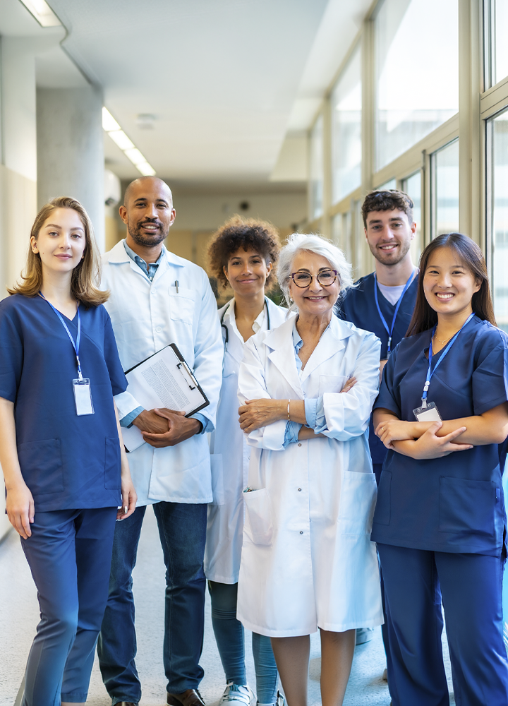 Grupo de profesionales sanitarios posando en el pasillo de un centro de salud u hospital. Varias personas llevan uniformes clínicos o batas blancas y están situadas frente a grandes ventanales con luz natural.