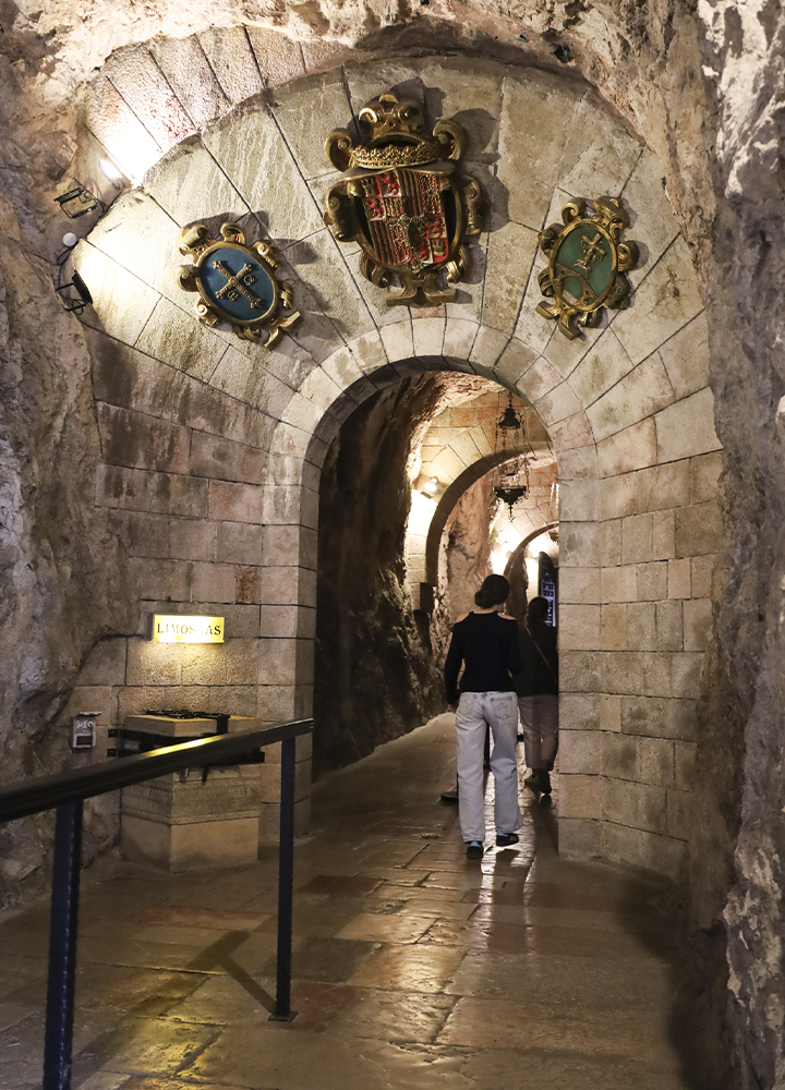 Pasadizo de piedra con arcos de mampostería y suelo empedrado, iluminado por luces cálidas. Sobre el arco principal cuelgan tres escudos decorativos. Dos personas caminan por el corredor, que se adentra en una galería subterránea.