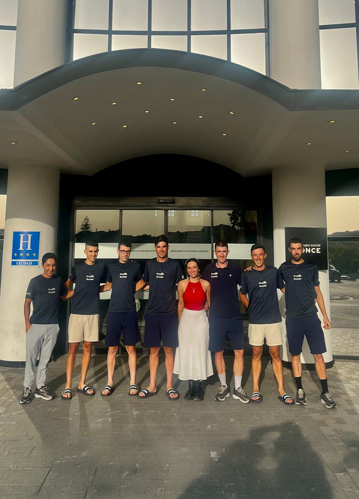 Grupo de personas posando frente a la entrada de un hotel, bajo una marquesina y junto a la puerta principal. El edificio presenta grandes columnas y ventanales, mientras varias personas visten equipación deportiva oscura. A un lado se aprecia la placa de categoría hotelera y un rótulo de la ONCE.
