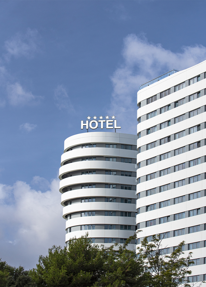 Edificio hotelero moderno de color blanco con fachada curva y varias plantas. En la parte superior destaca el rótulo “HOTEL” acompañado de cinco estrellas. El edificio se recorta sobre un cielo azul con algunas nubes blancas y vegetación en la parte inferior de la imagen.
